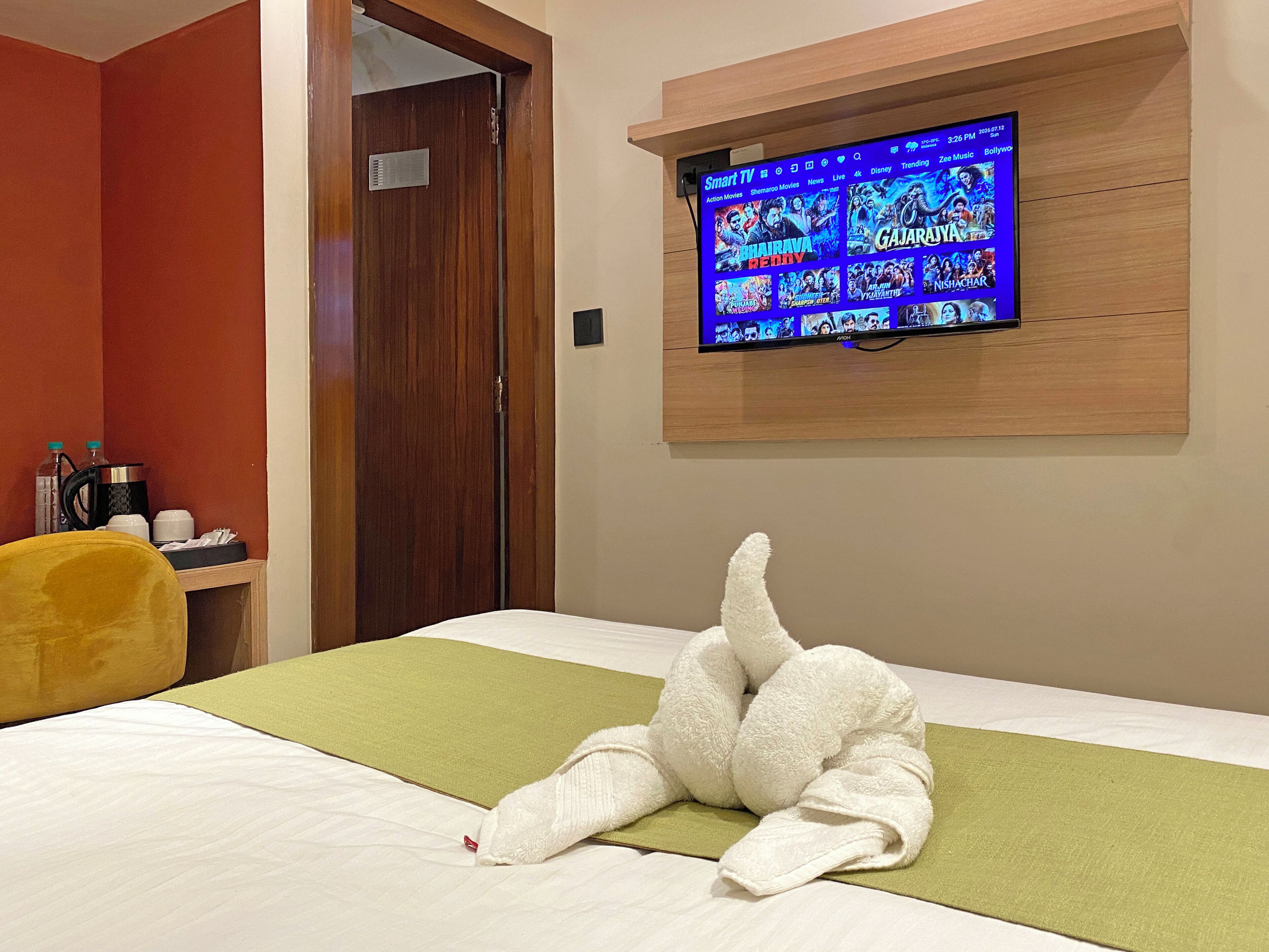 Habitación de hotel con cama, televisor montado en la pared mostrando opciones de películas y puerta de madera.