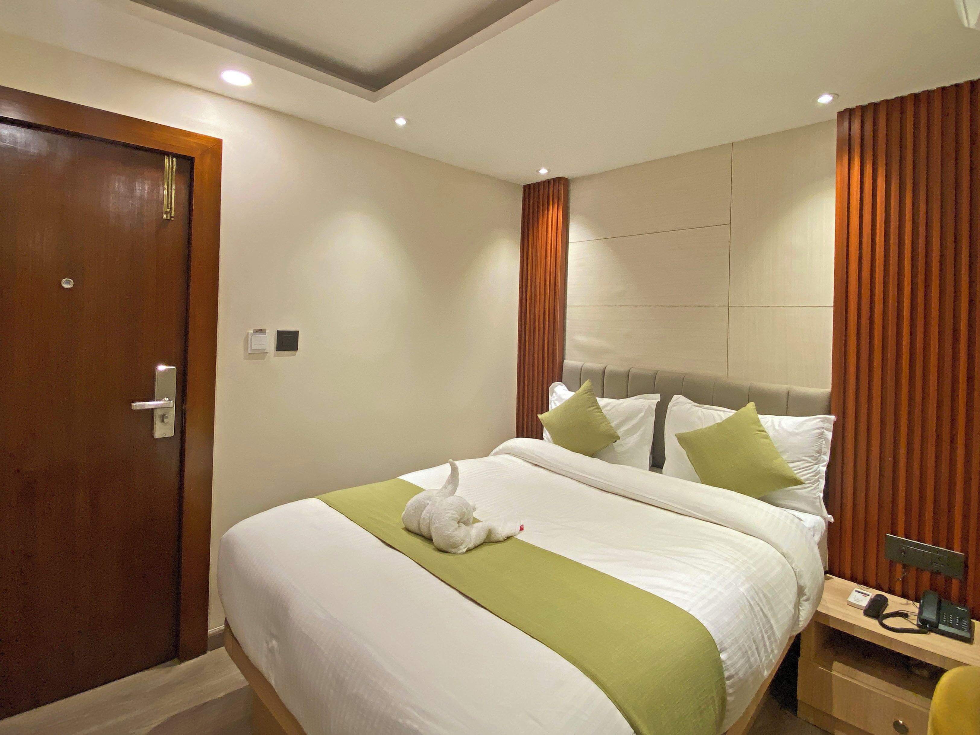 Habitación de hotel con cama, una puerta de madera, una mesita de noche con teléfono y un panel de control en la pared.