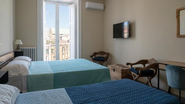 A hotel room with two beds, a television, a wooden chair, a desk, and a window with a view of buildings. - Bellavista Relais (Napoli)