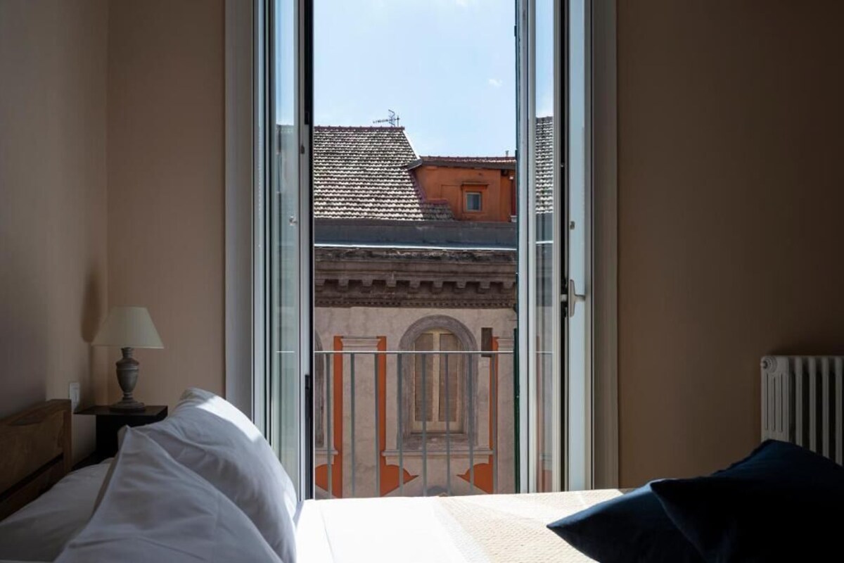 Ein Hotelzimmer mit einem Bett, einem Nachttisch mit Lampe und Blick auf Gebäude durch eine Glastür.