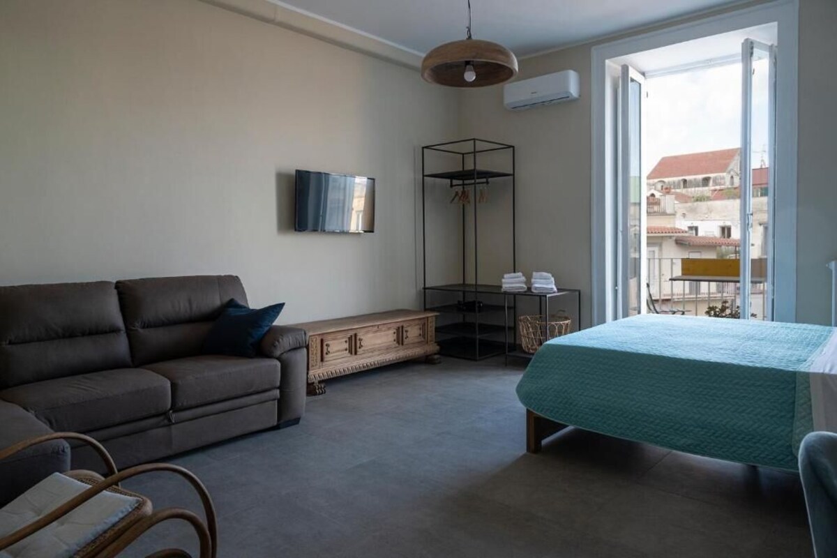 Ein Schlafzimmer mit Bett, einer Couch, einer Holzkommode, einem Fernseher und einem Balkon mit Blick auf Gebäude.