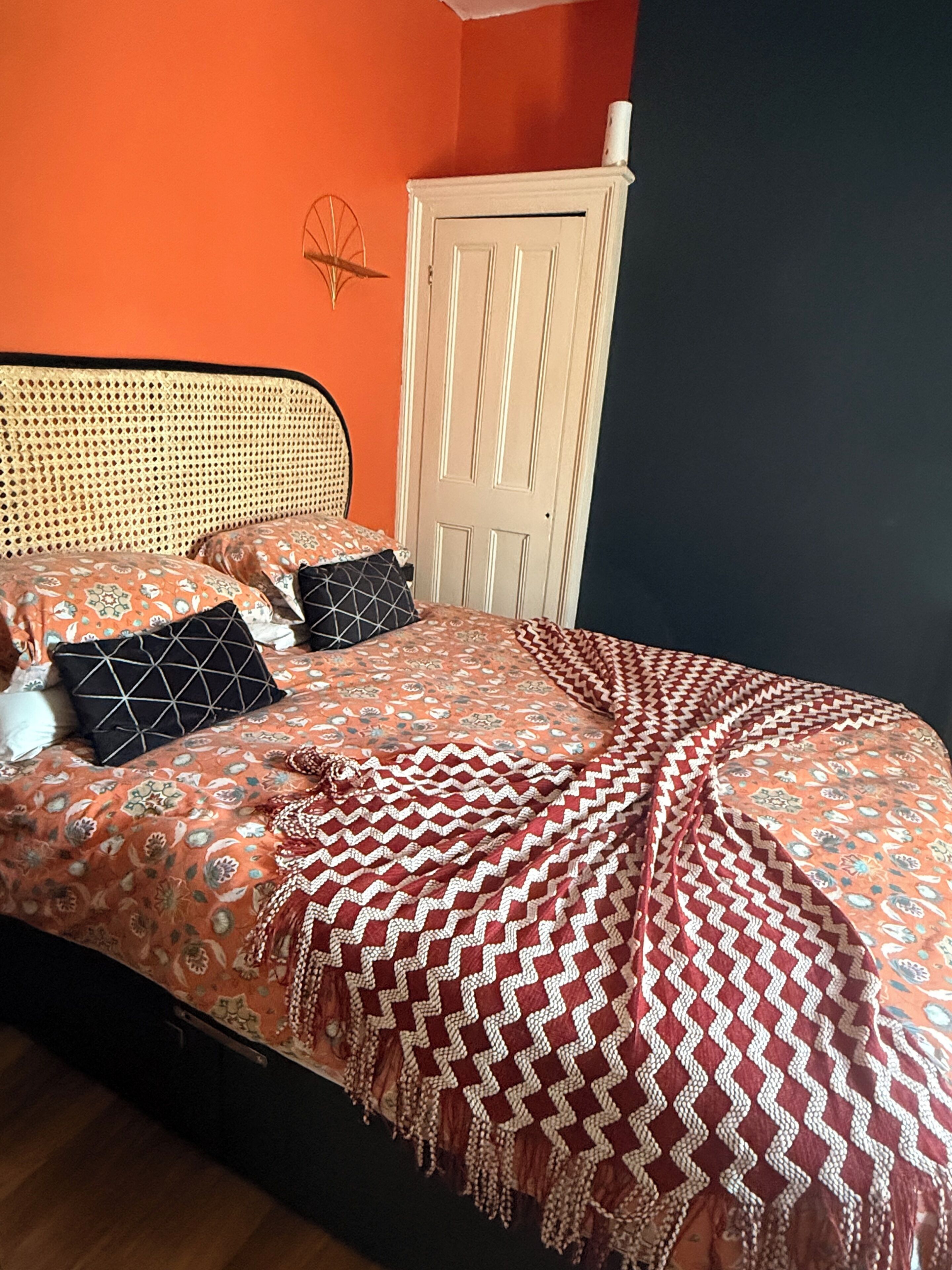 Una cama con colcha estampada y almohadas, un cabecero tejido y una puerta blanca con una toalla encima.