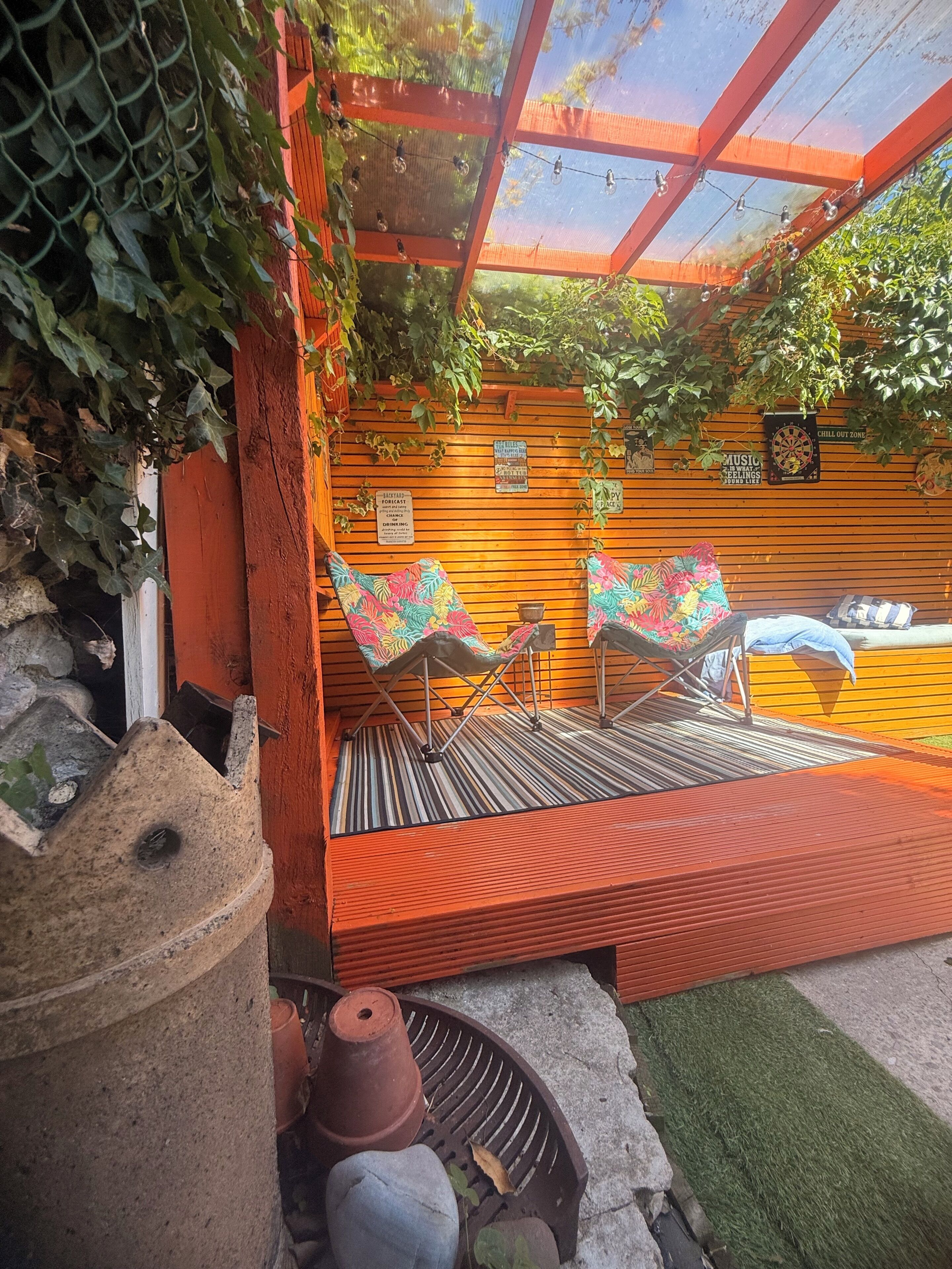 A covered outdoor seating area with a wooden bench, two chairs with colorful cushions, and a small table.