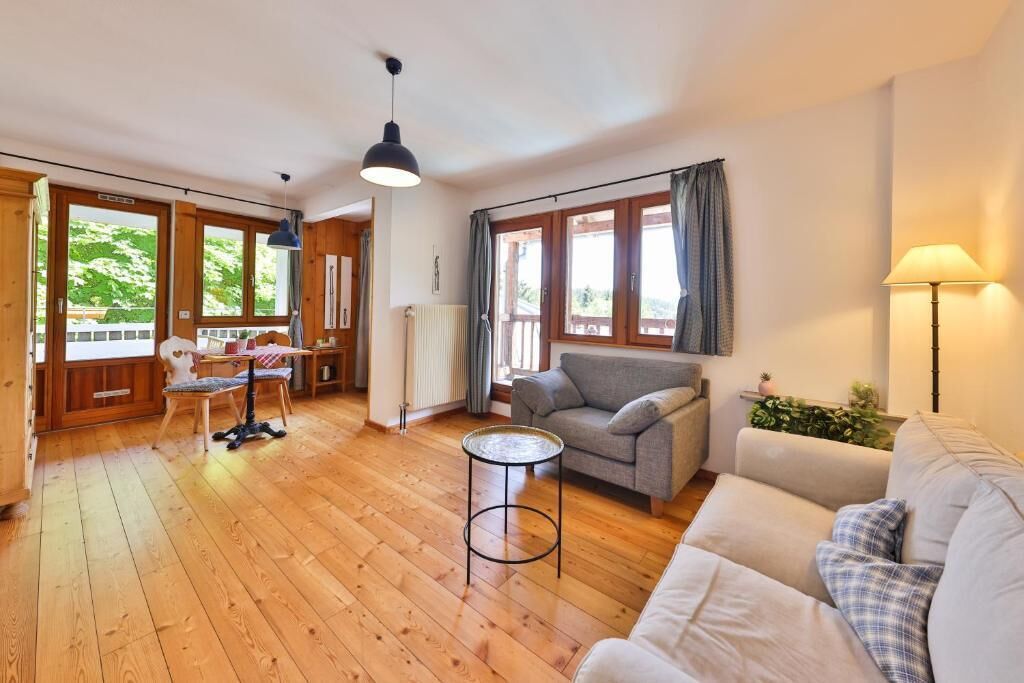 A spacious living area with wooden flooring, a sofa, a table, and a chair. There are large windows with curtains, a lamp, and a radiator.