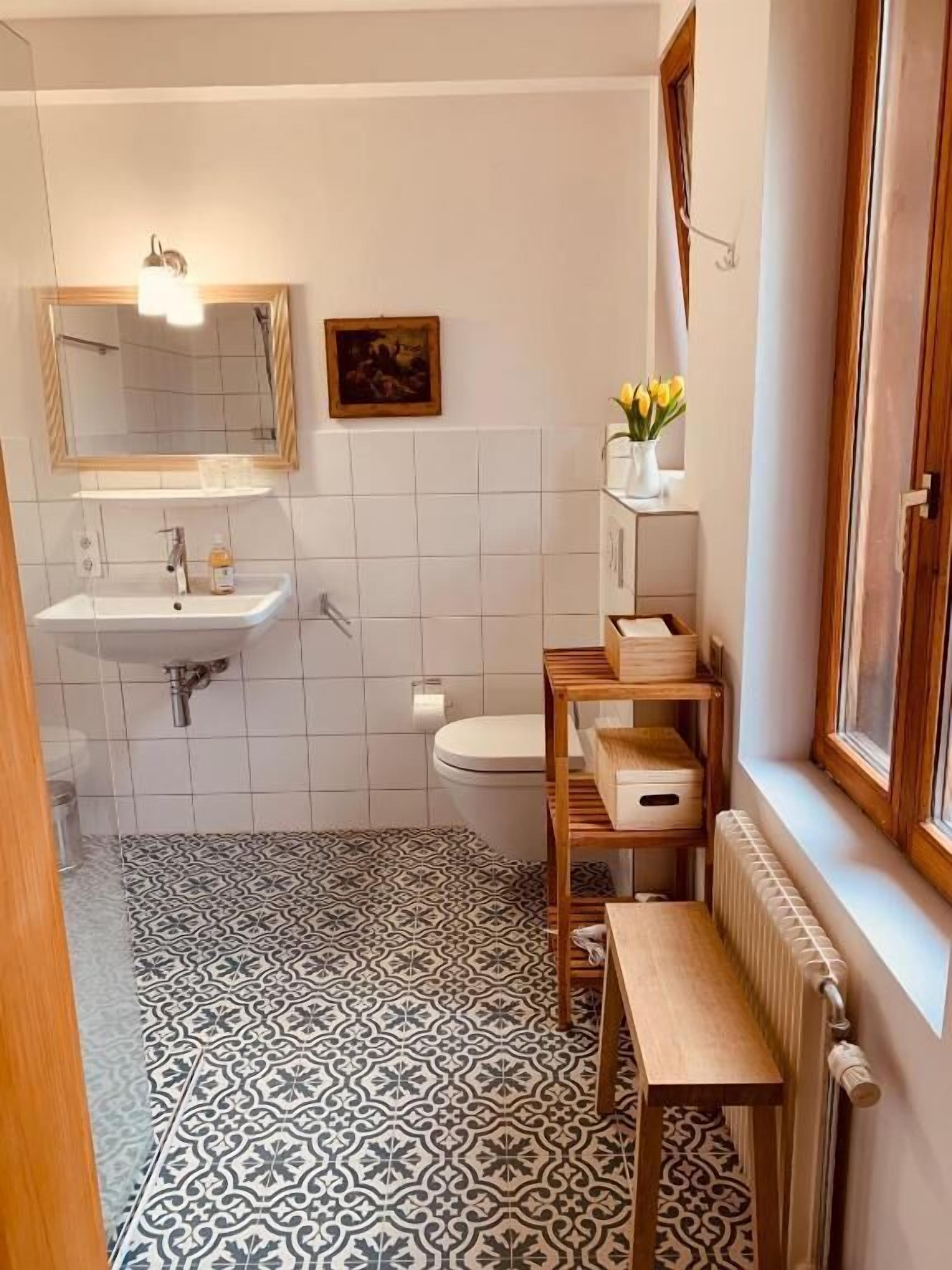 Ein Badezimmer mit einem weißen Waschbecken, einem Spiegel, einer Toilette, gefliestem Boden mit Muster, einem Heizkörper und einem Fenster mit Vorhängen.