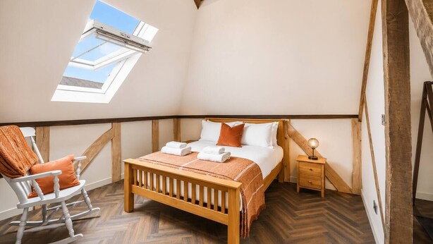Un dormitorio con techo inclinado, tragaluz, cama de madera, una mecedora y una mesita de noche con lámpara.