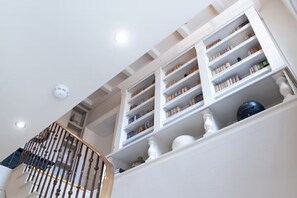 Ein mehrgeschossiges Interieur mit weiß gestrichenen Wänden, einer Treppe mit Holzbalkon und einer Reihe von Regalen, die mit unterschiedlichen Gegenständen gefüllt sind.