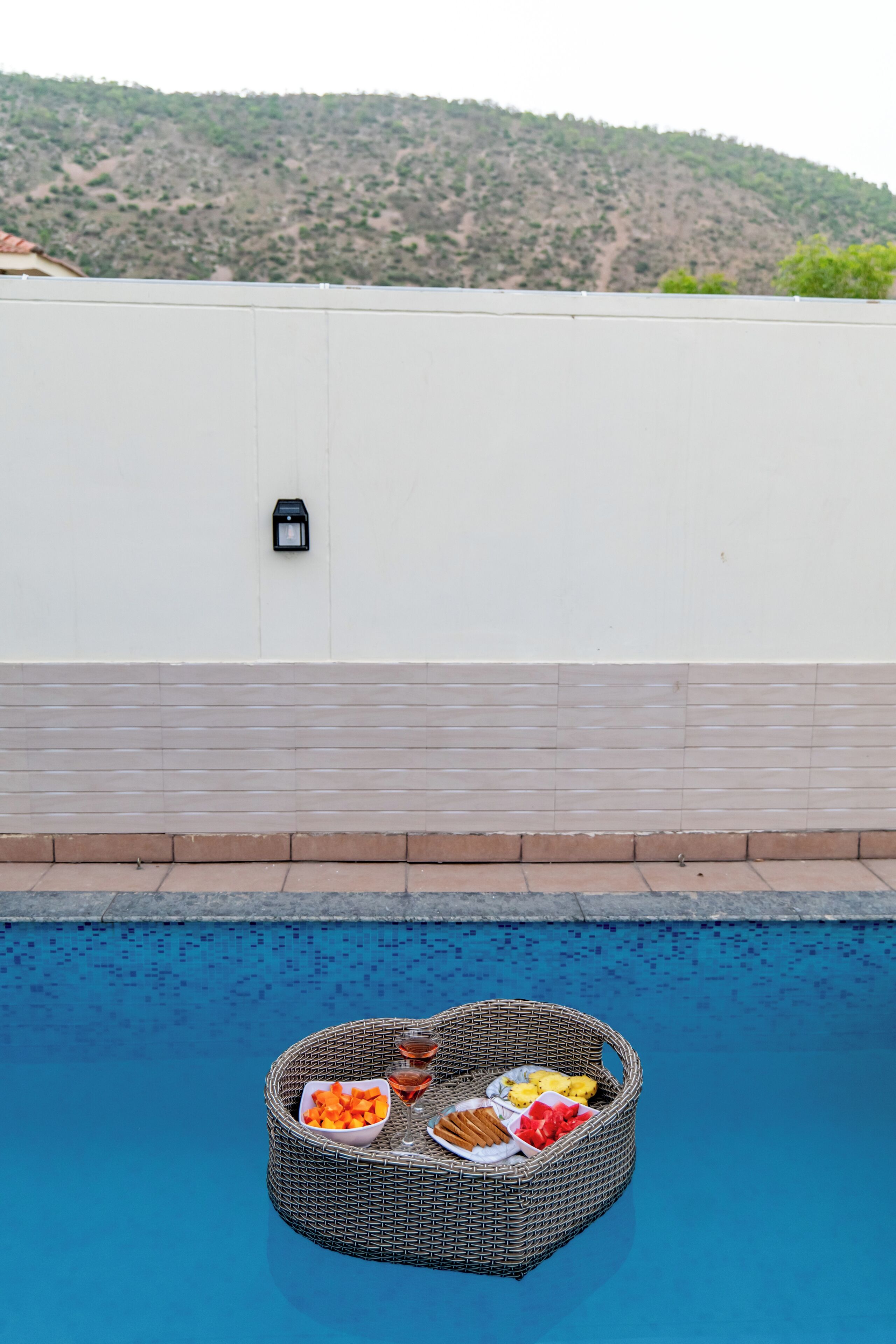A poolside area with a wicker lounge chair filled with snacks and drinks, a tiled pool, and a white wall with a small black object.