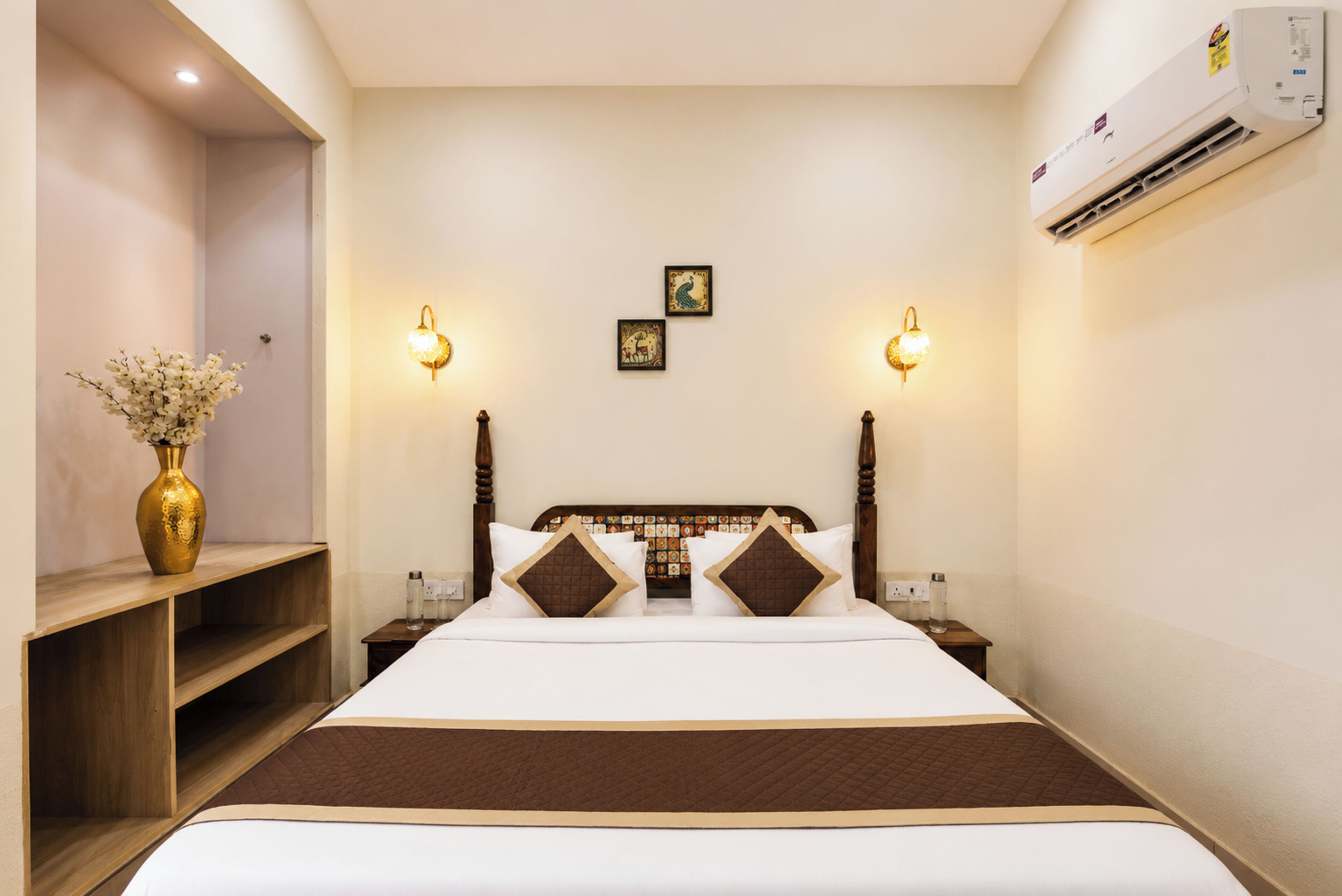 A hotel room with a large bed, bedside tables, a vase of flowers, wall-mounted lights, and an air conditioning unit.