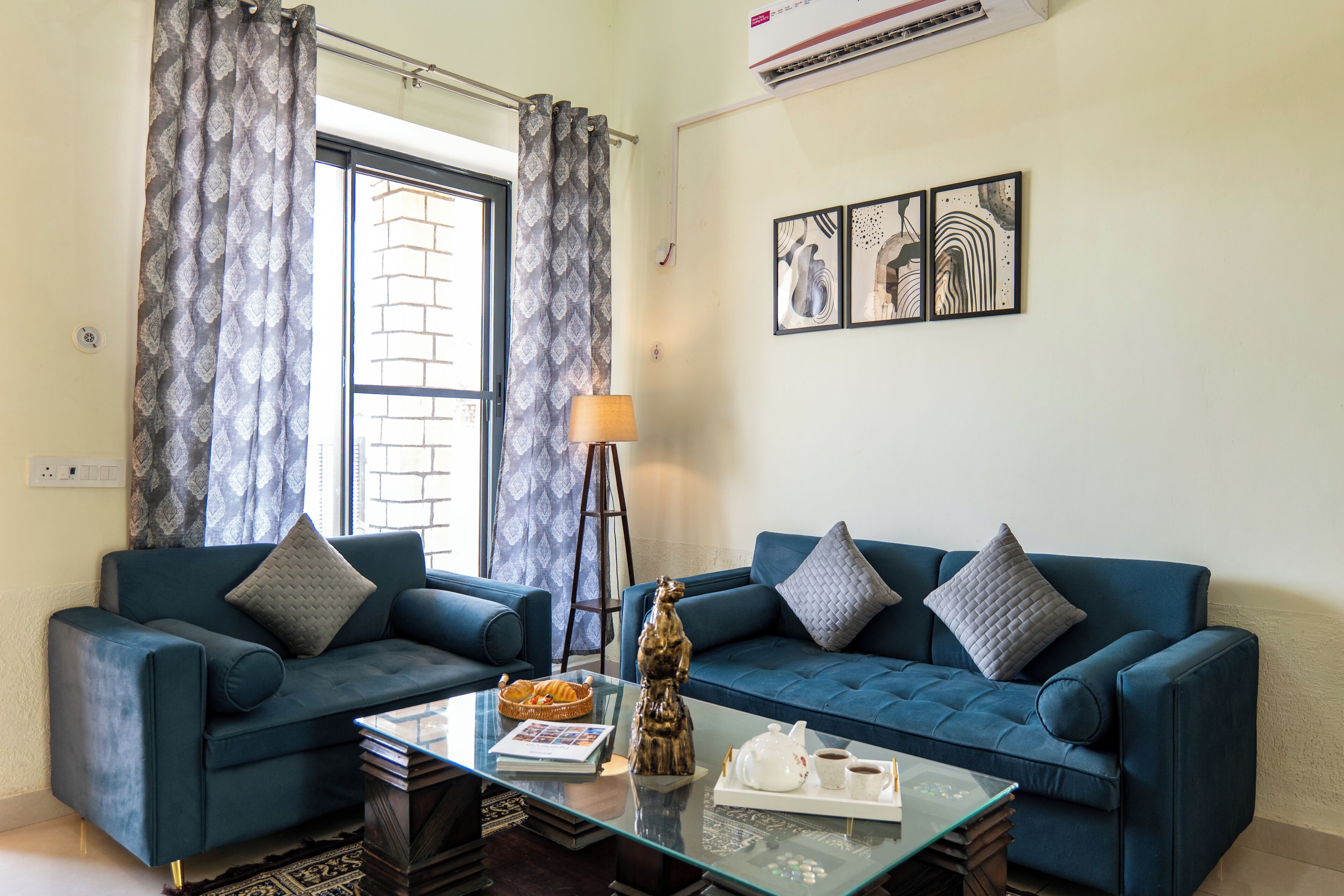 A living room with two blue sofas, a glass coffee table, a floor lamp, and a wall with abstract art.