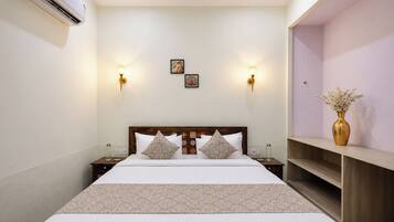 A hotel room with a large bed, two bedside tables, a nightstand, a wall-mounted air conditioner, two wall sconces, and a small shelf with a vase.