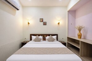 Habitación de hotel con una cama grande, dos mesitas de noche, un tocador, aire acondicionado montado en la pared, dos apliques y un pequeño estante con un florero.
