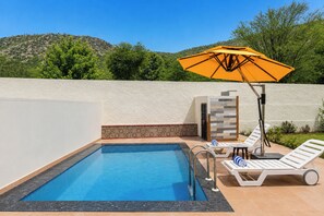 Una piscina con solárium de baldosas, sillas de descanso, una mesa con un vaso y una sombrilla.