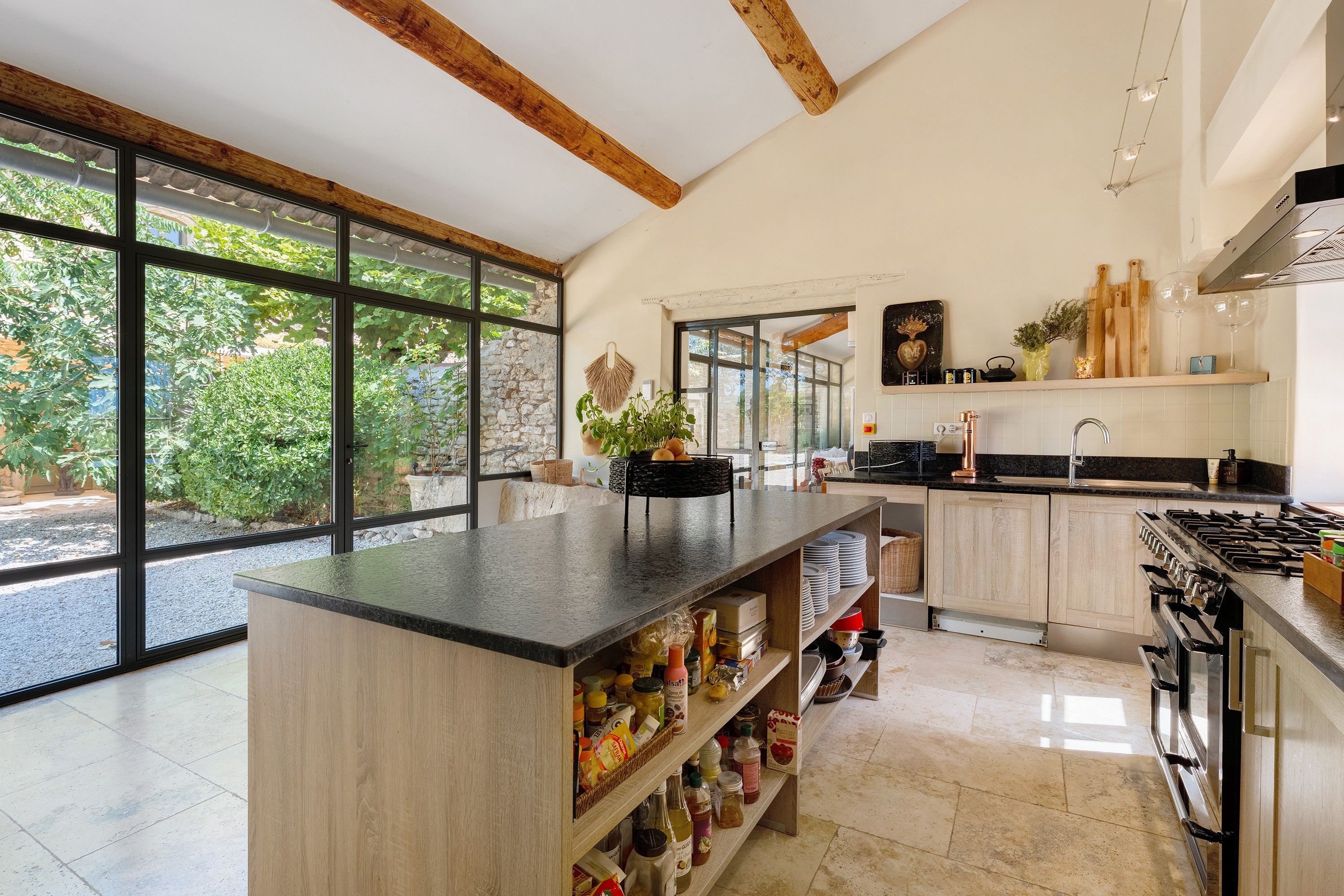 A modern kitchen with a central island, large windows, and a view of greenery outside.