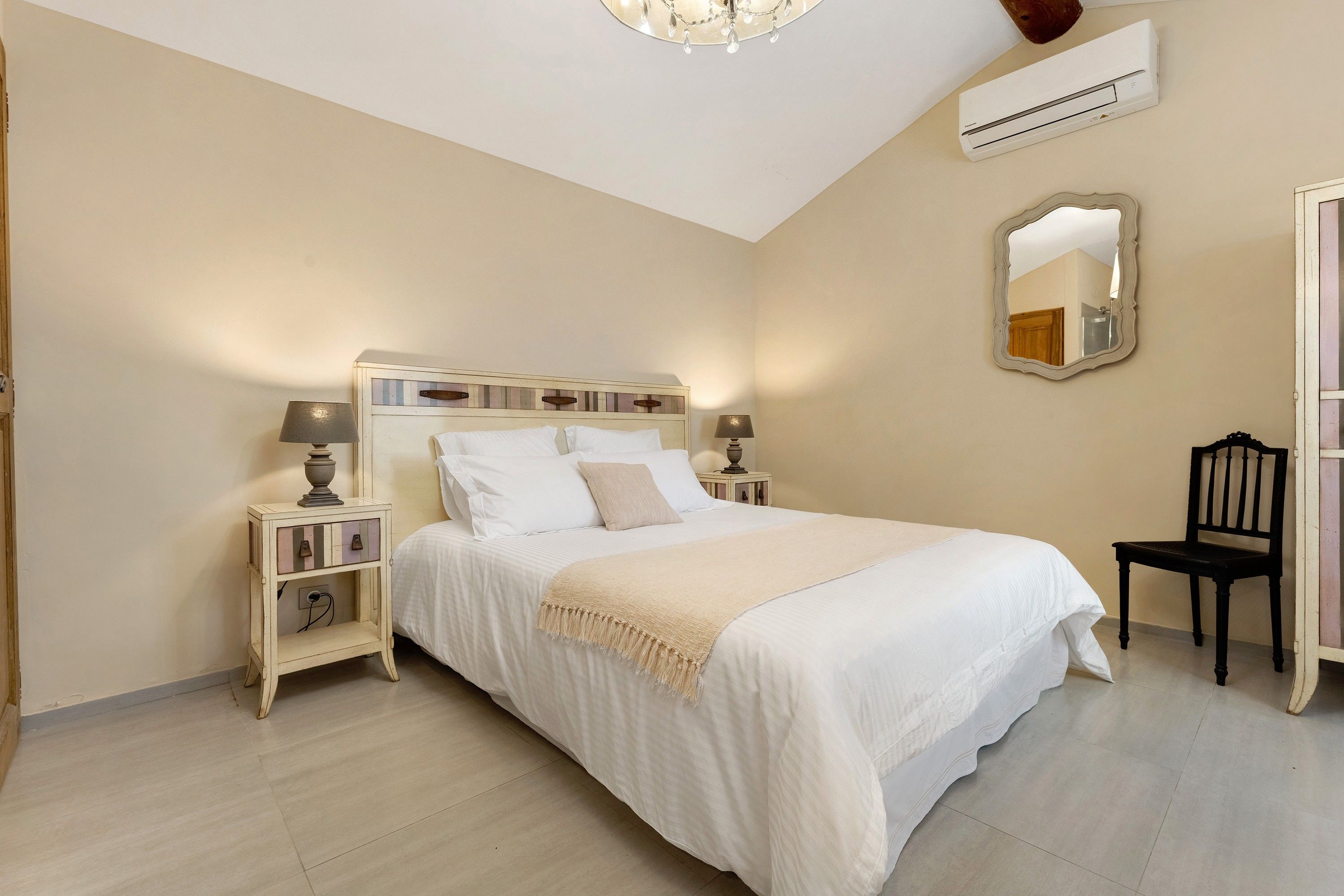 Ein Schlafzimmer mit einem Bett, Nachttischen, einem Sessel, einem Spiegel und einer Klimaanlage.