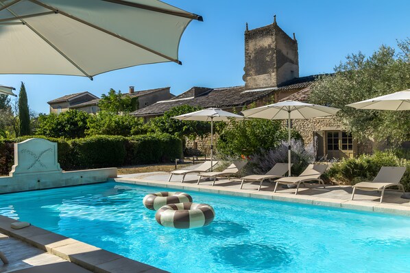 Piscina con sillones de playa y sombrillas, rodeada de vegetación y un edificio de piedra con una torre.