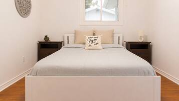 A neatly made bed with a light-colored bedspread and a pillow with text, two bedside tables with lamps, a small potted plant, and a window with a view of the outside.