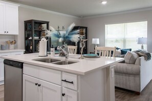 A modern kitchen with white cabinetry, a central island with a sink, and a dining area with a chair and table. - New Palm Coast Stay | Golf, Parks & Beach (Palm Coast)