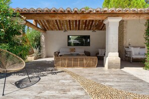 Un espacio de estar al aire libre techado, con un banco tejido, una silla de mimbre y un pilar de piedra.