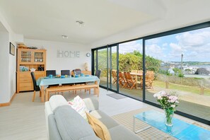 A modern living space with a dining area, a sofa, a glass coffee table, and a view of the outdoors. - House With Grand Harbor Views (Falmouth)