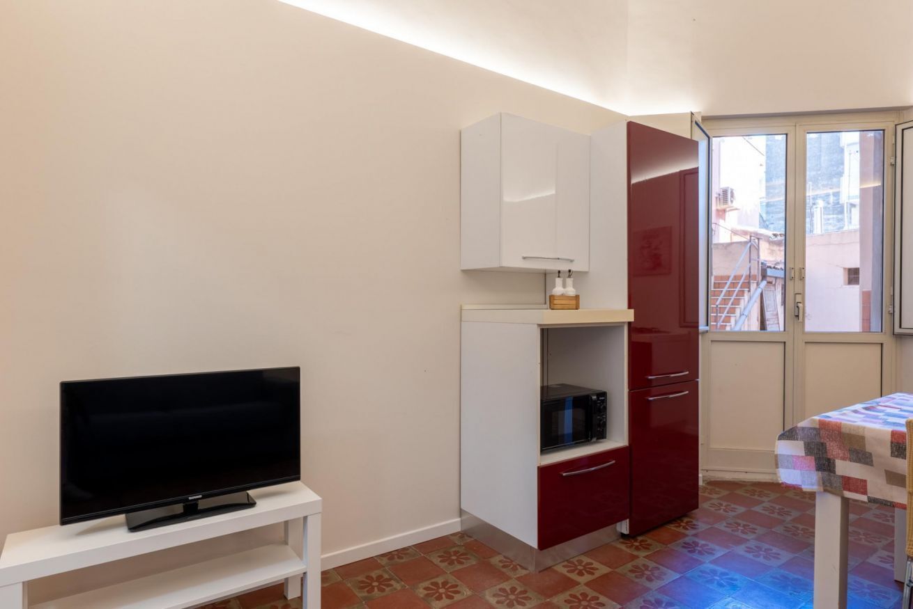 Een kleine kamer met een televisie op een witte standaard, een magnetron op een rode kast en een tafel met een tafelkleed met patroon.