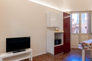 Ein kleiner Raum mit einem Fernseher auf einem weißen Schrank, einer Mikrowelle auf einem roten Korpus und einem Tisch mit gemustertem Tischläufer.