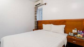 A hotel room with a wooden headboard, a bed with white bedding, a nightstand with a phone and a basket of items, and an air conditioning unit on the wall.