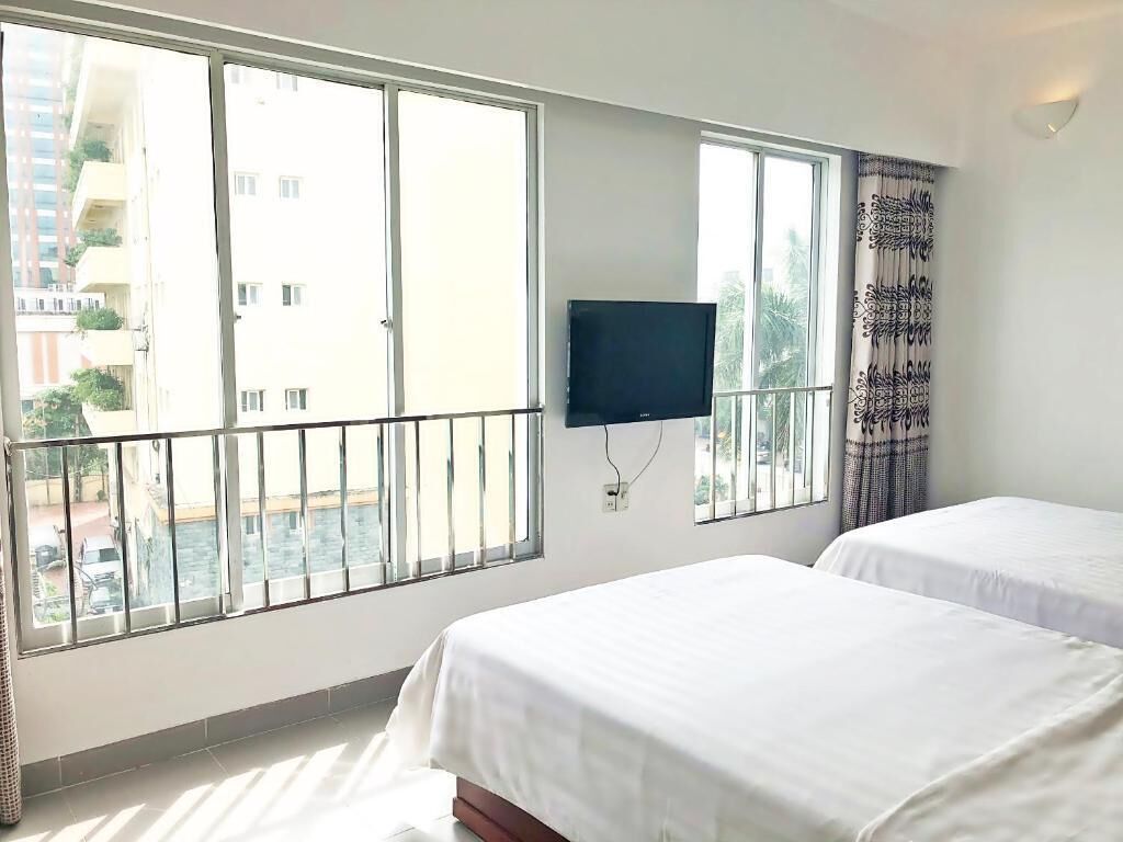 Ein Hotelzimmer mit zwei Betten, einem Fernseher und großen Fenstern mit Blick auf Gebäude und Grünflächen.