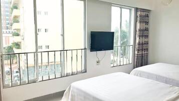 Ein Hotelzimmer mit zwei Betten, einem Fernseher und großen Fenstern mit Blick auf Gebäude und Grünflächen.