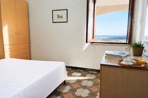 Ein Zimmer mit einem Bett, einem Holzschrank, einem gerahmten Bild an der Wand, einem Fenster mit Meerblick und einem Tisch mit Frühstücksgebäck.