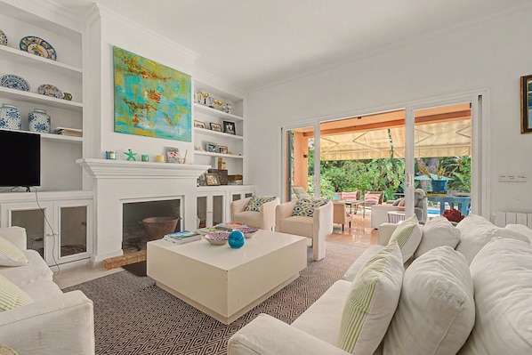 A living room with a fireplace, white sofas, a coffee table, and a large window with an outdoor view.
