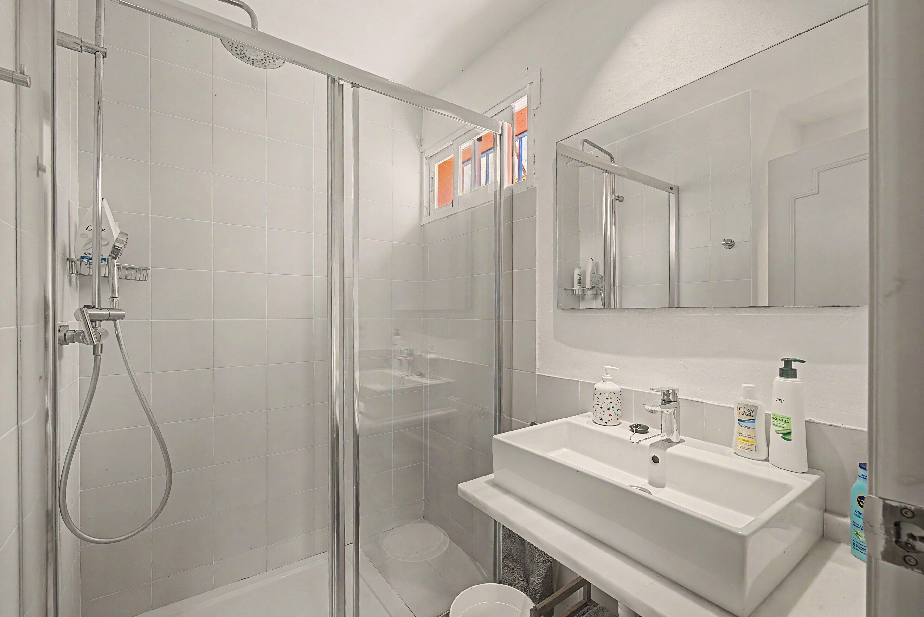 A modern bathroom with a glass-enclosed shower, a white sink with a mirror above, and toiletries on the countertop.