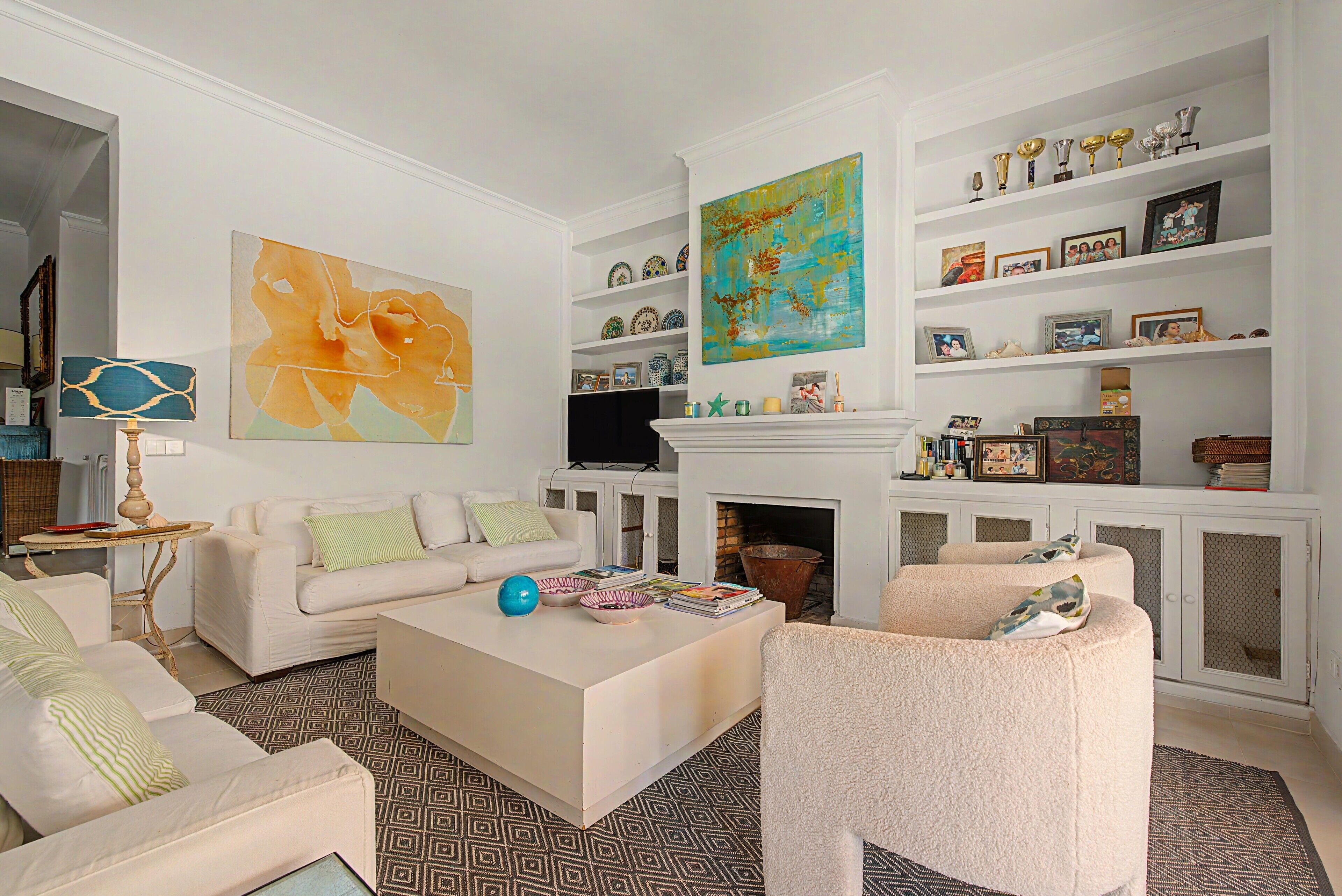 A living room with a white sofa, a coffee table, and a fireplace. There are shelves with decorative items and a large abstract painting.