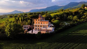 A large, historic building with multiple levels and a prominent tower, surrounded by lush green fields and mountains in the background.