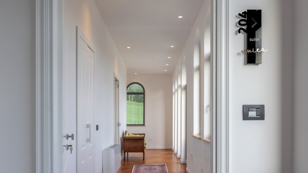 A hallway with white walls, a wooden floor, and a rug leading to a room with a window. - RELAIS FLORA BERTA (Matelica)