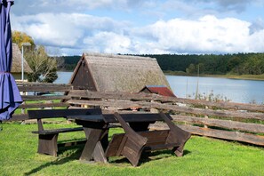 A rustic wooden structure with a thatched roof, situated by a lake with a clear sky above.