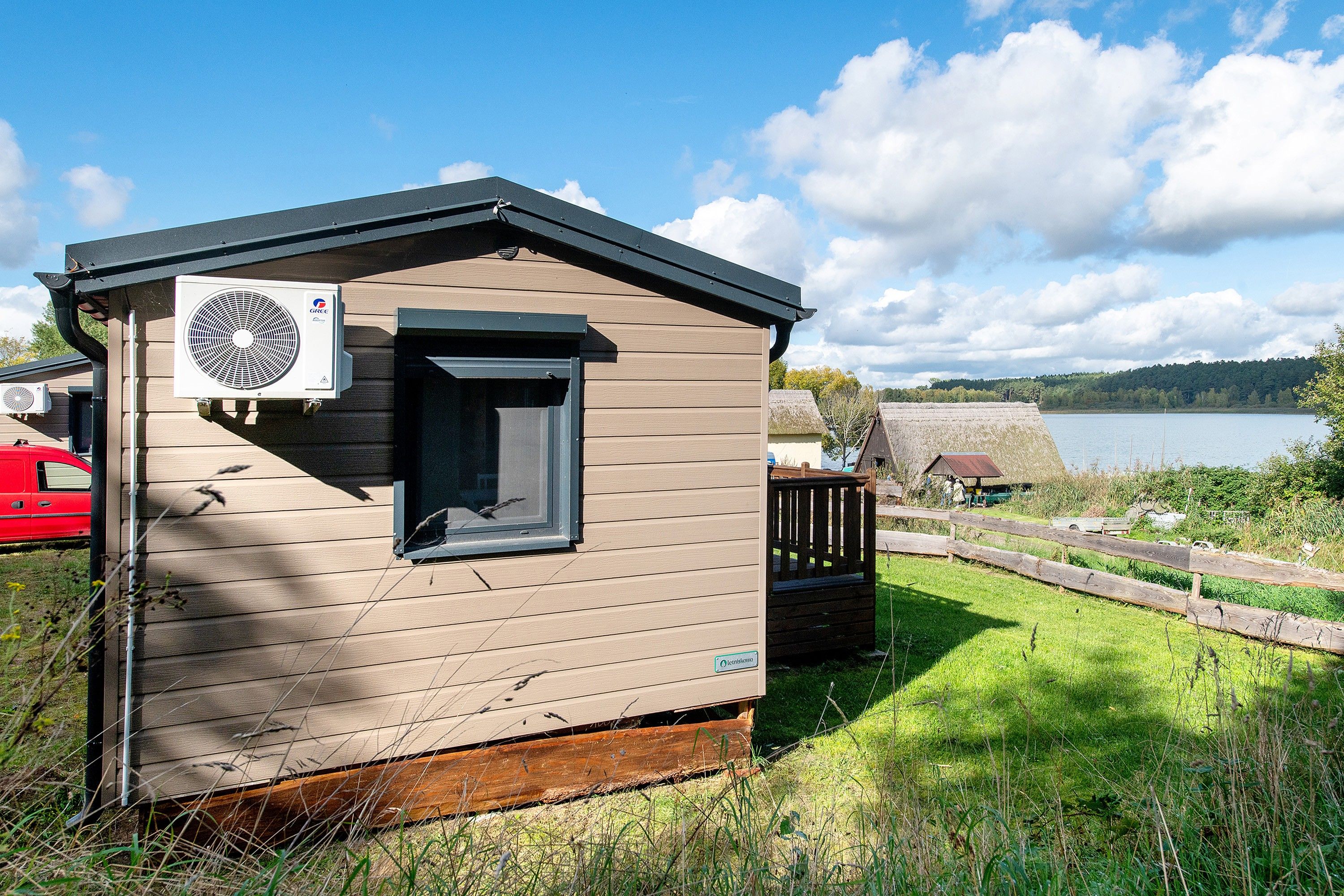 Eine kleine Holzhütte mit Fenster und Klimaanlage, in der Nähe von Wasser, mit Holzzaun und einem roten Fahrzeug im Hintergrund.