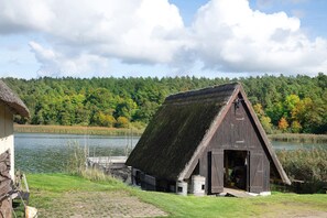 A rustic wooden building with a steep roof, situated by a calm body of water, surrounded by lush greenery and a dense forest in the background.