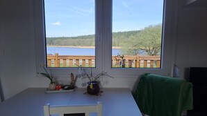 A room with a table and chairs, a window with a view of a lake and a wooden deck.