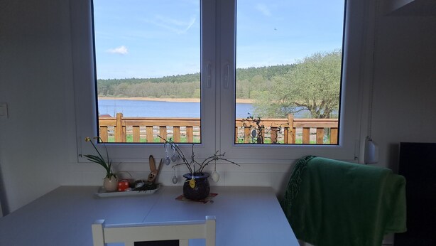 A room with a table and chairs, a window with a view of a lake and a wooden deck. - Tiny Home 'Haus Barsch' with Lake View, Wi-Fi and Air Conditioning (Lohmen)