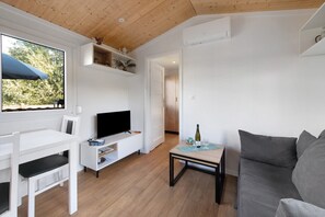 A modern living room with a television, a sofa, a small table with a bottle and glasses, and a window with a view of trees.