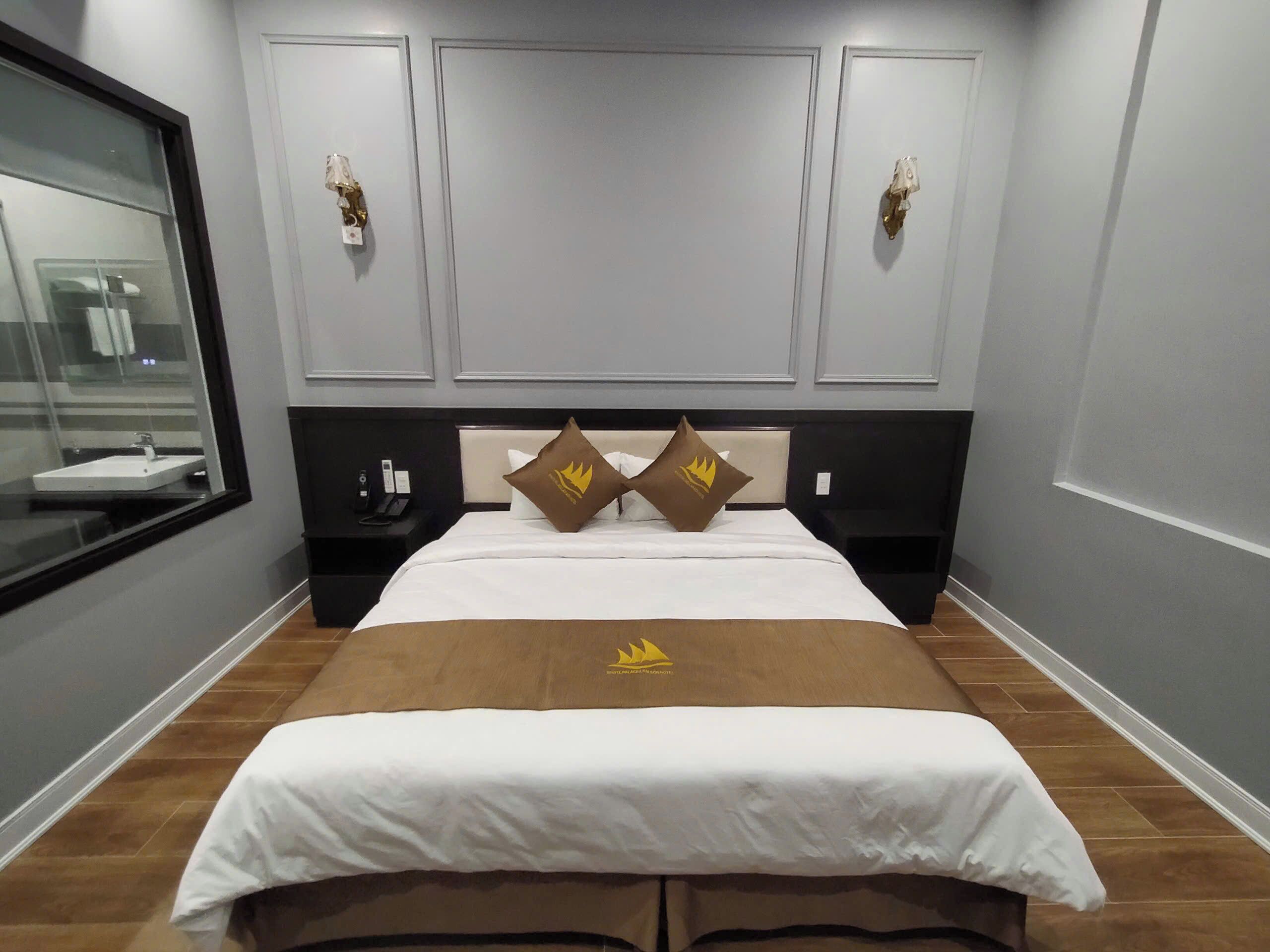 A hotel room with a bed, a bedside table, a nightstand, a window, and a wall-mounted light fixture.