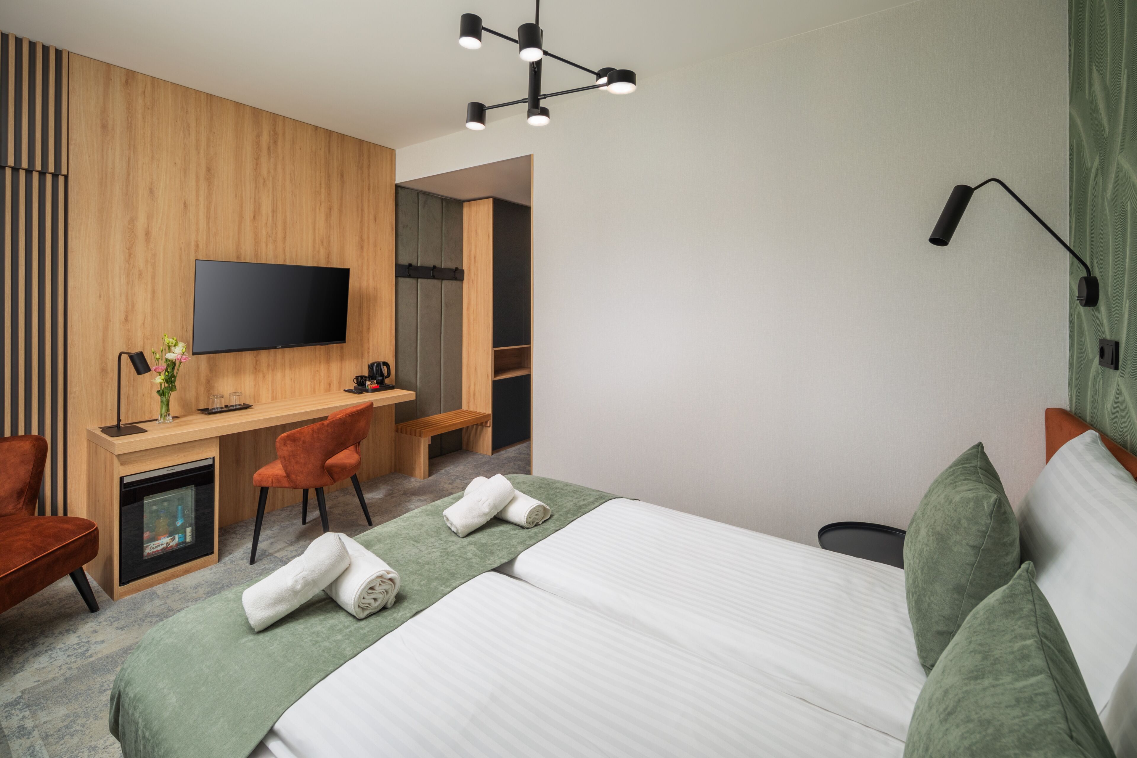 A modern hotel room with a large bed, a desk with a chair, a flat-screen TV mounted on the wall, and a small seating area with a bench.