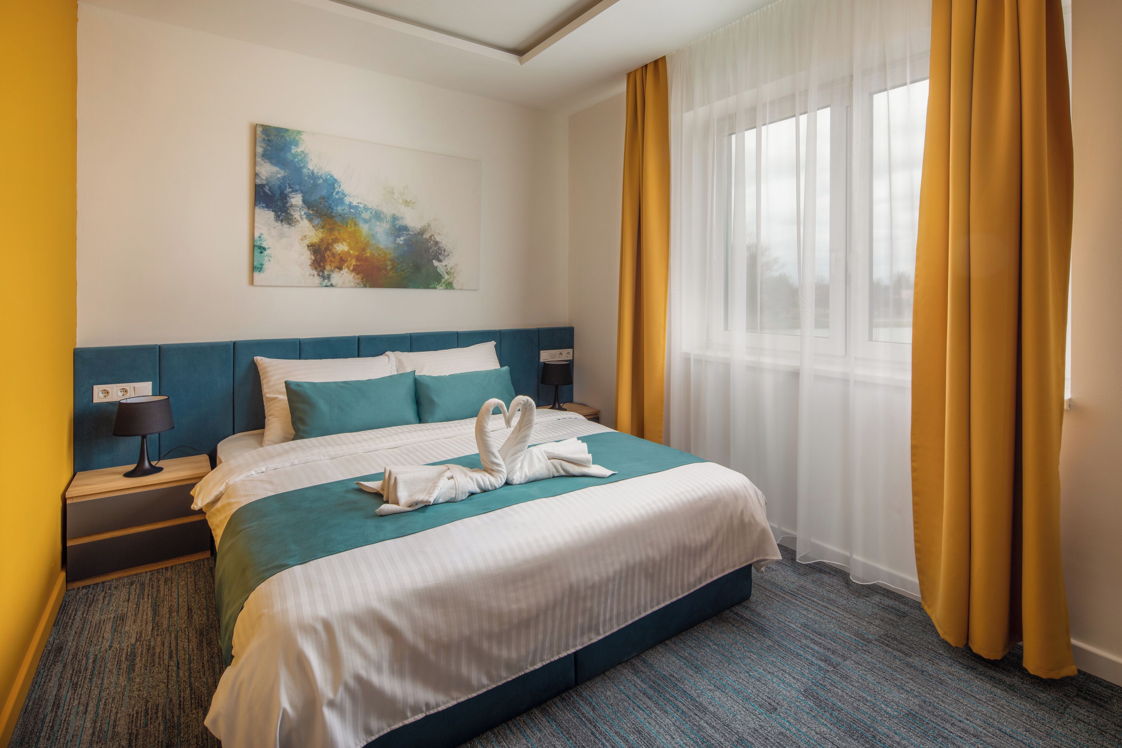 Une chambre d’hôtel avec un grand lit, une table de chevet, une lampe, un tableau au mur et une fenêtre avec des rideaux.