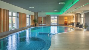 A large indoor swimming pool with a wooden deck surrounding it, large windows, and a view of the outside. - Sziget Hotel (Nyíregyháza)