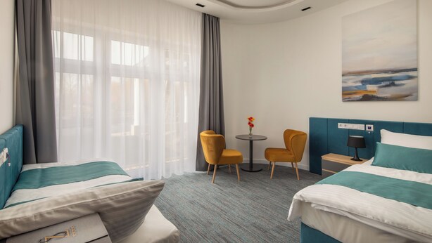 A hotel room with two beds, a small table with chairs, a painting on the wall, and a view of the outdoors through the window. - Sziget Hotel (Nyíregyháza)
