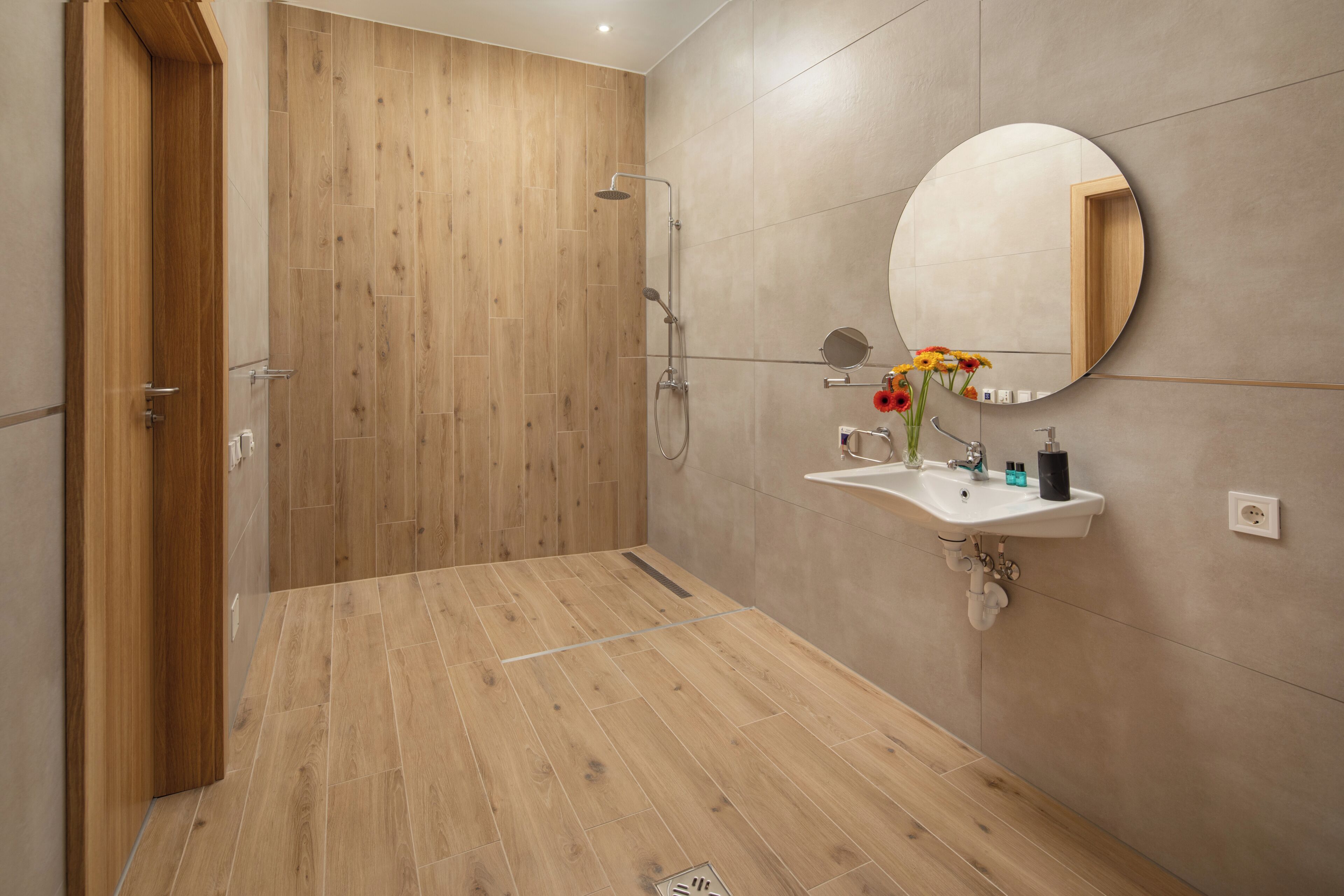 A modern bathroom with a wooden shower enclosure, a round mirror, and a sink with a vase of flowers.