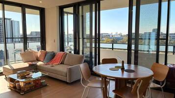 A modern living room with a sofa, coffee table, and dining table. Large windows offer a view of the cityscape.