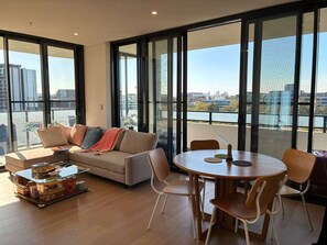 A modern living room with a sofa, coffee table, and dining table. Large windows offer a view of the cityscape. - New & Quiet 2BR/2BA with Sydney Tower Views, Close to Metro & Shopping Centre (Macquarie Park)