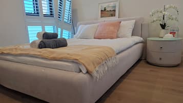 A modern bedroom with a bed, bedside tables, a window with blinds, and a painting on the wall.